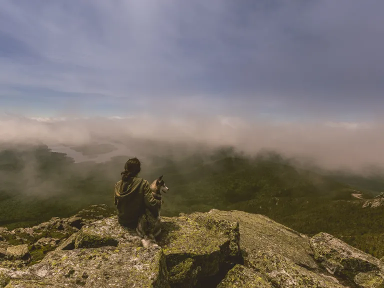 Even when we have unanswered prayers, know that God does listen. A woman sitting on cliff edge with her dog.